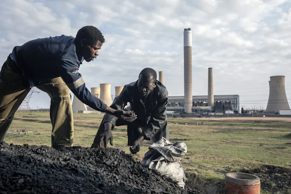 Two men put coal chunks into a sack with a power plant in the background.