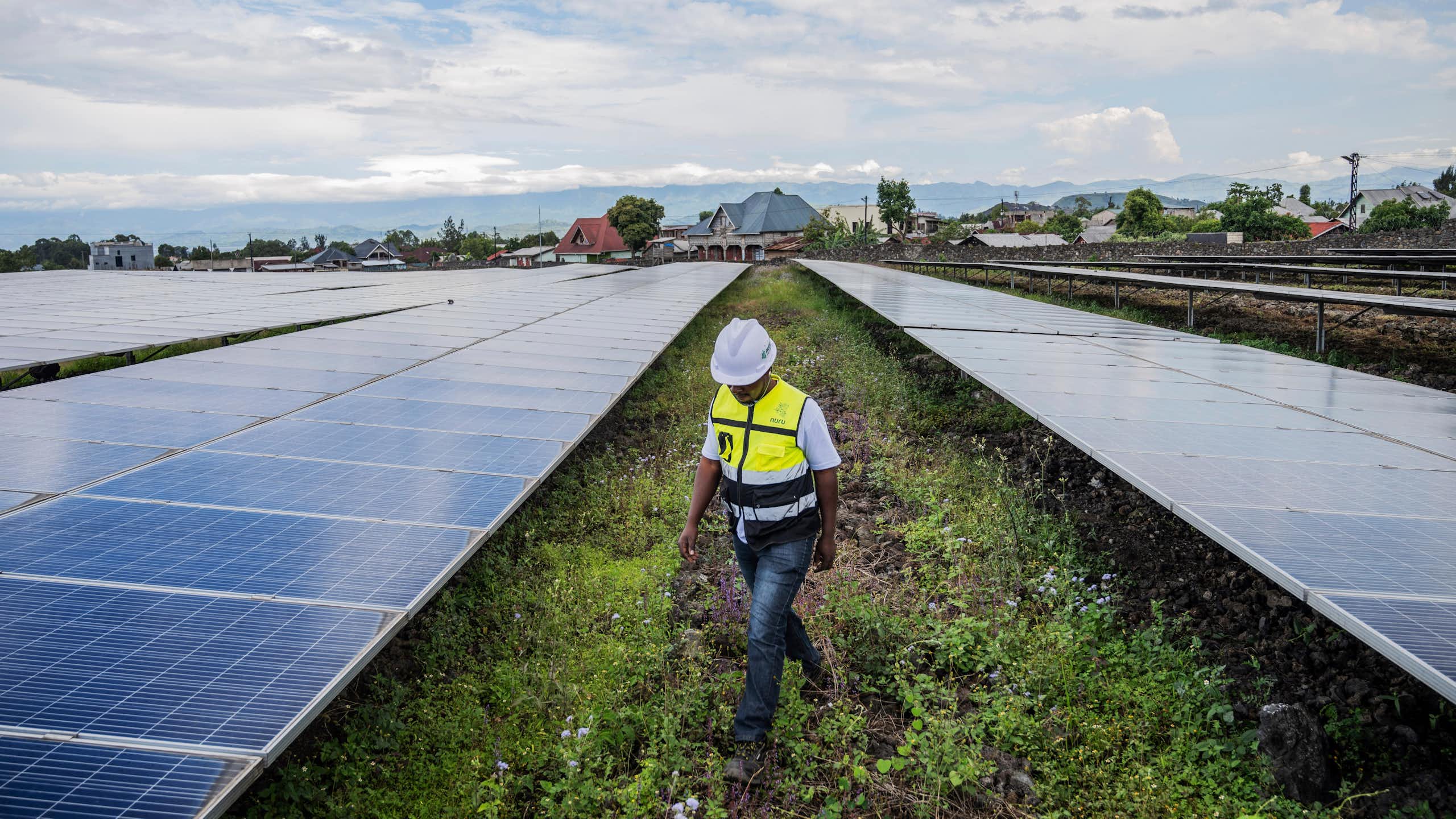 A worker in a hard hat walks between rows of solar panels. Farms are in the backgorund.