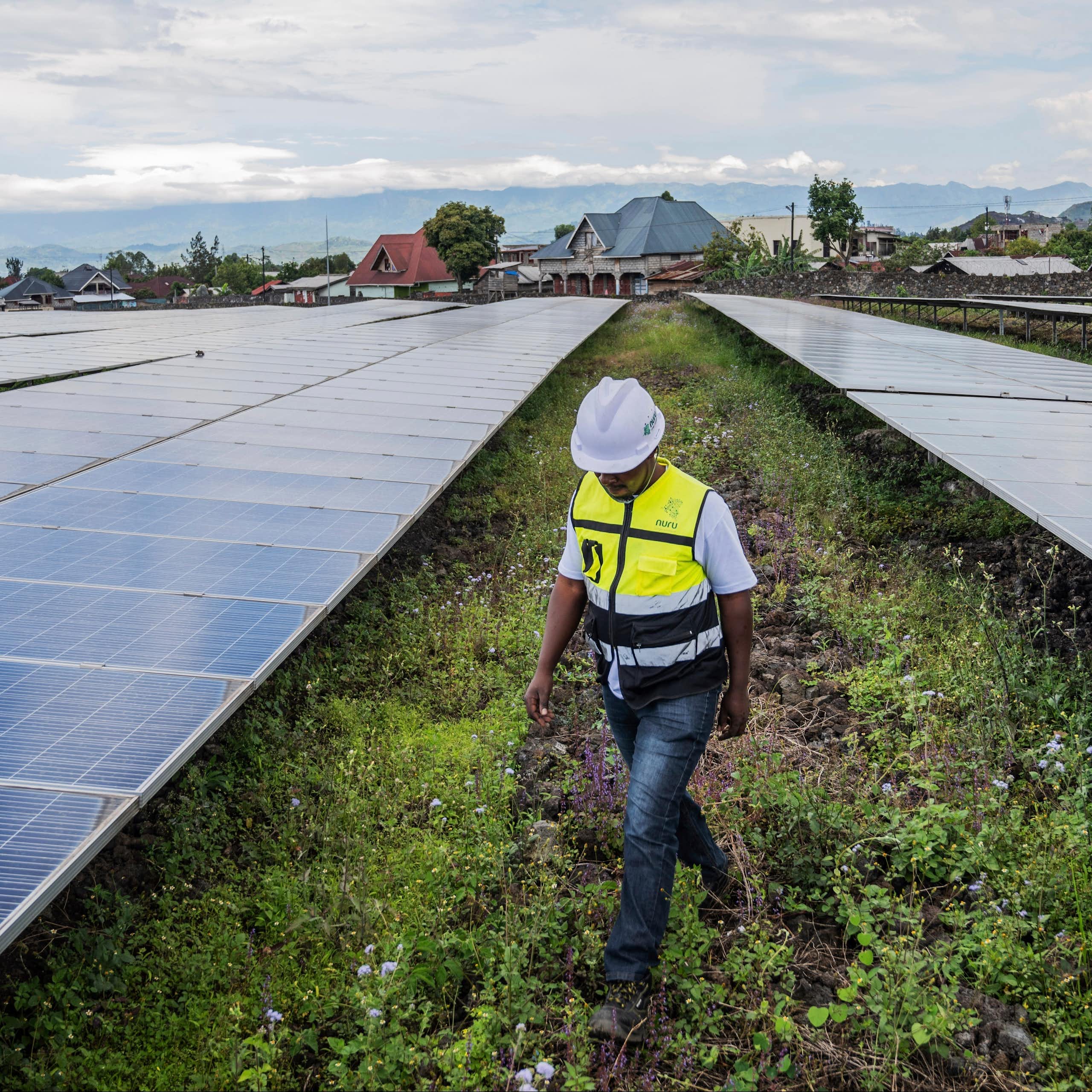 A worker in a hard hat walks between rows of solar panels. Farms are in the backgorund.