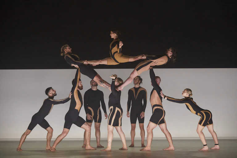A group of contemporary dancers in black and tan bodysuits create a human sculpture on stage, with three lifted by the others.