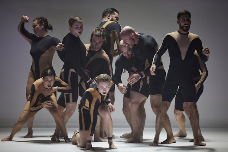 A group of contemporary dancers in black and tan bodysuits pose in a tight cluster on a bare stage, each in a slightly different pose.