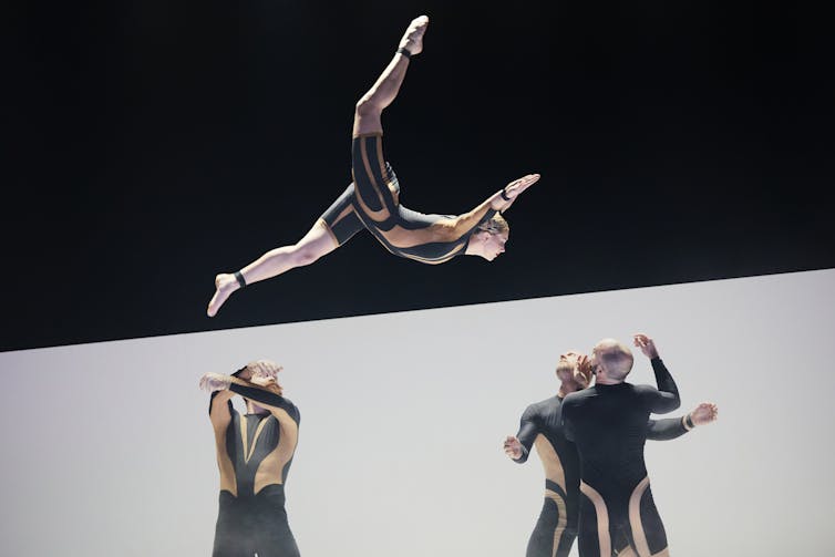 A dancer is launched high into the air, fully extended in a split-like position with arms reaching forward. Below, three other dancers in matching costumes look up as if preparing to catch him.