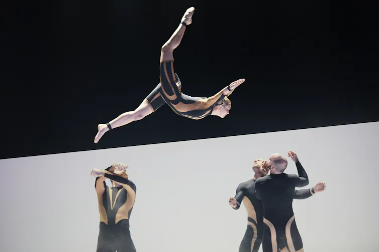 A dancer is launched high into the air, fully extended in a split-like position with arms reaching forward. Below, three other dancers in matching costumes look up as if preparing to catch him.