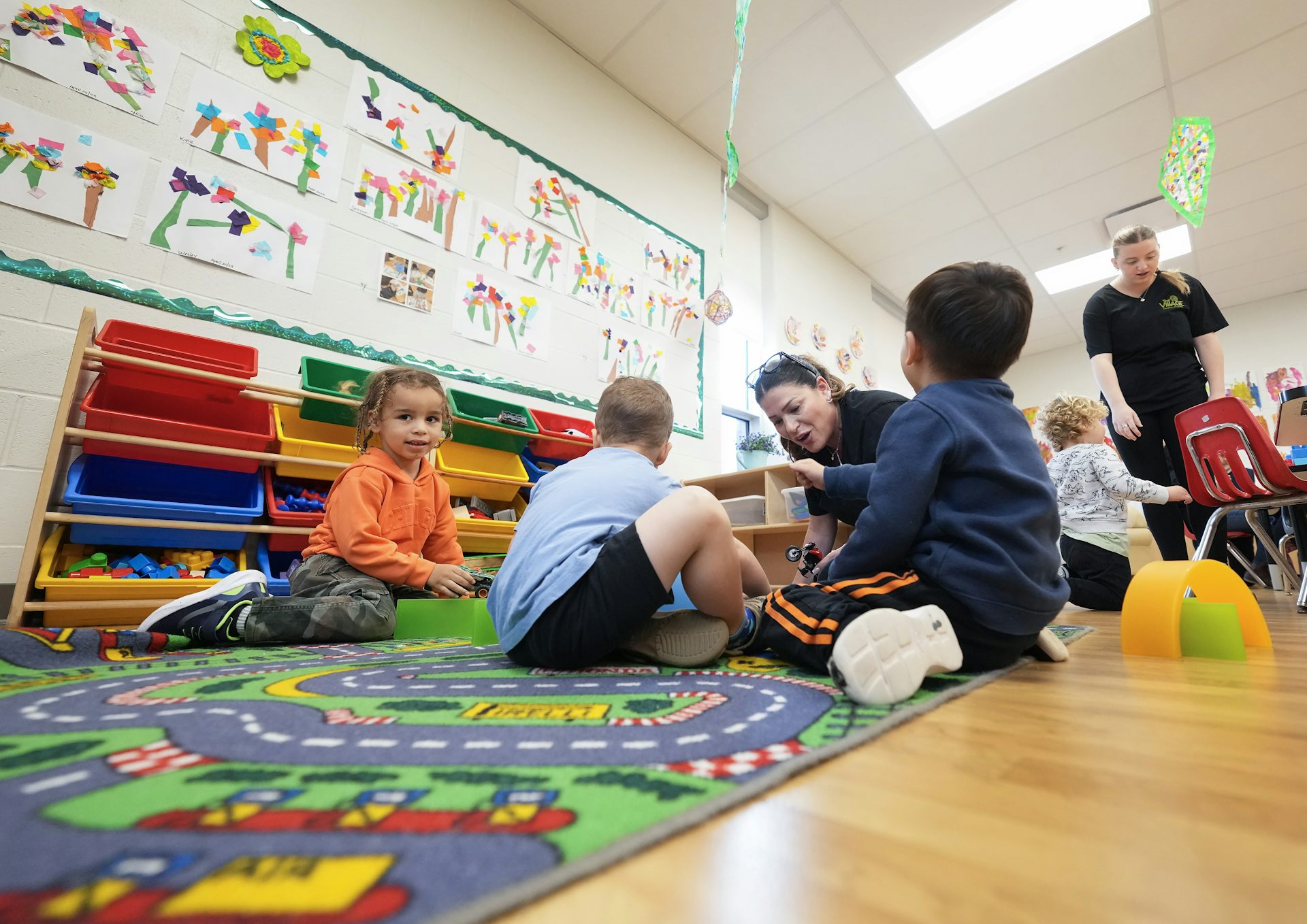 Children with toys on a carpet while adlts are nearby.