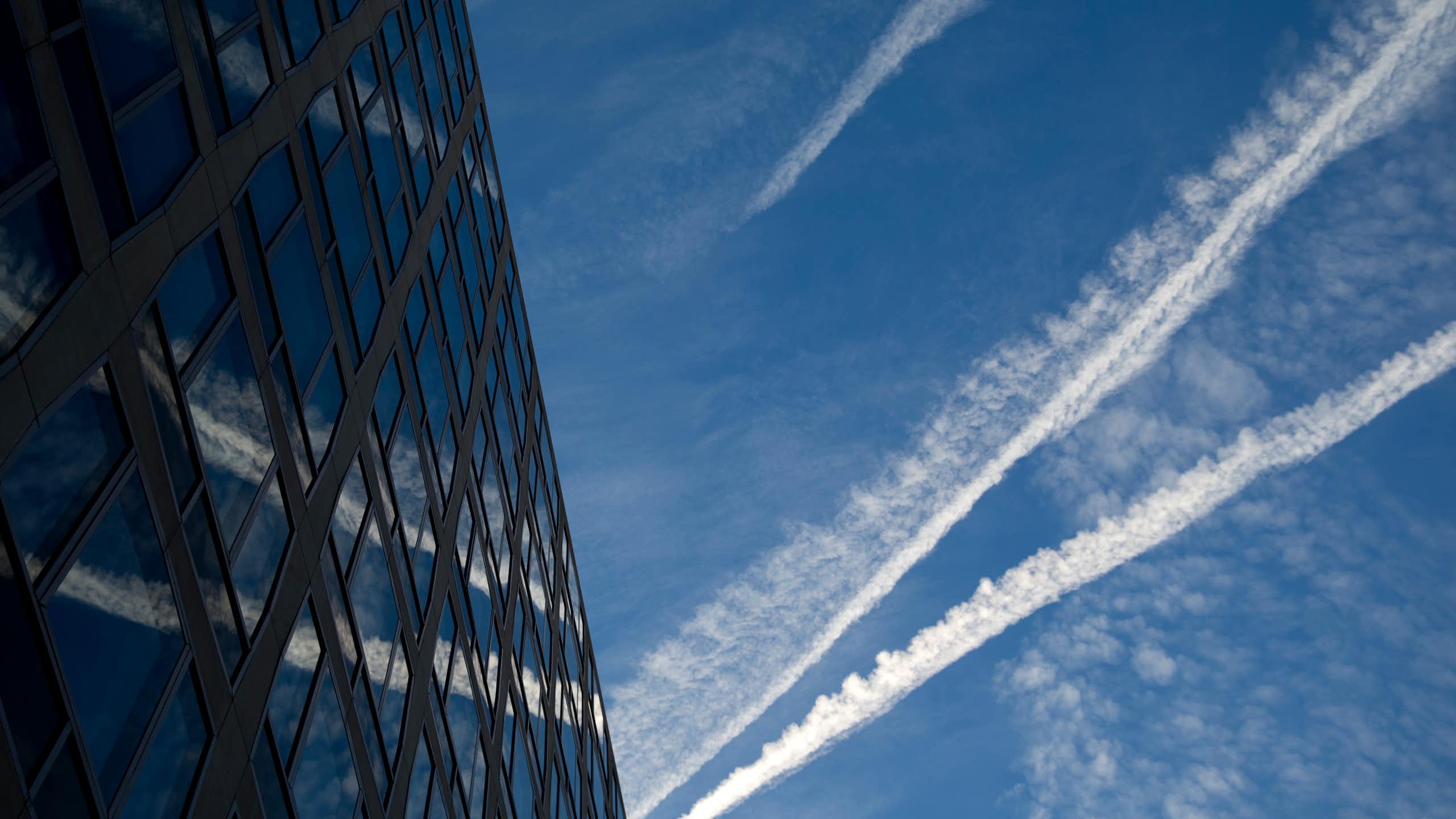 contrails against a blue sky and reflected in a large glass-front building