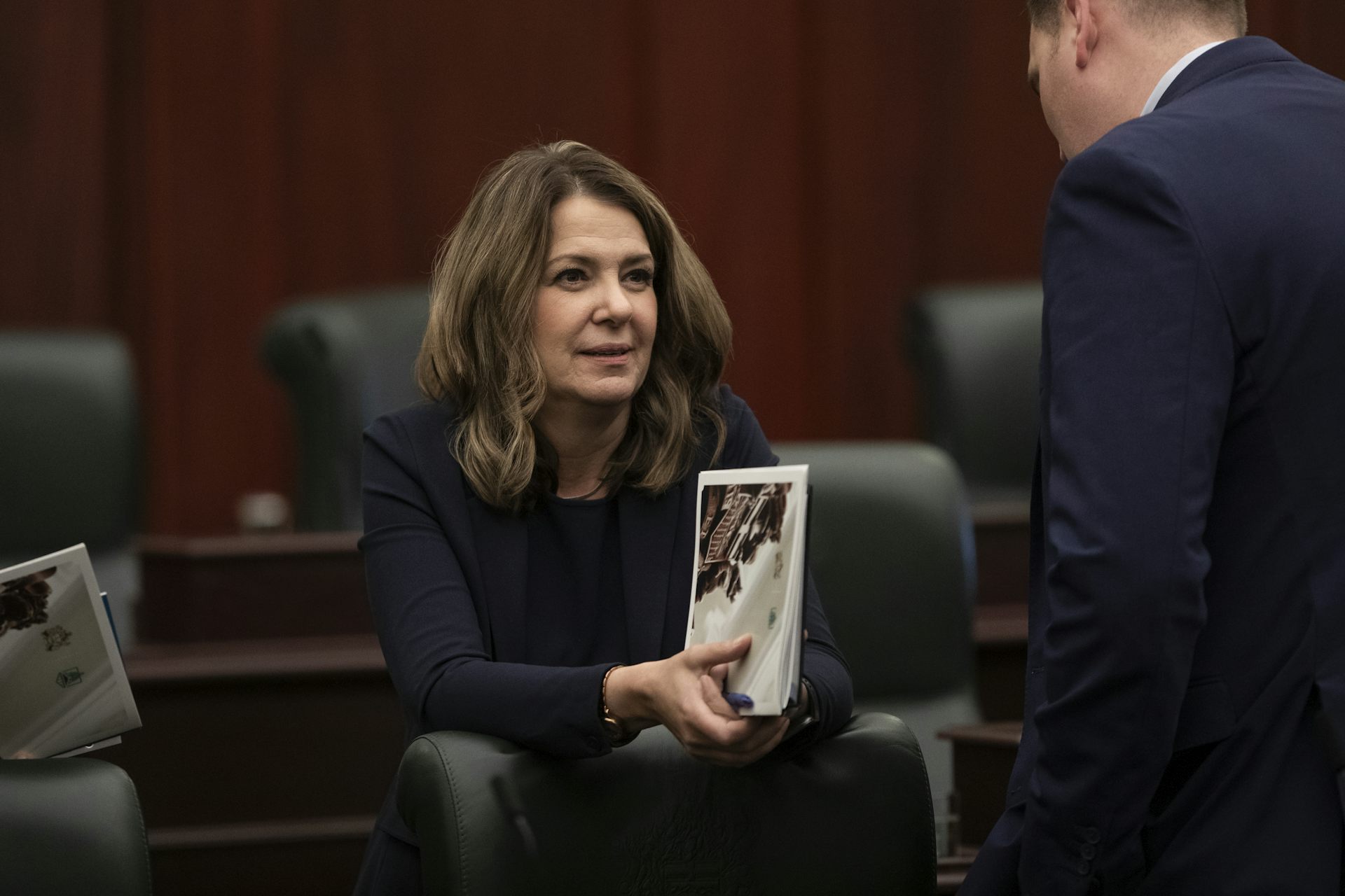 A person holding a binder seen leaning across a table in a space of rows of chairs.