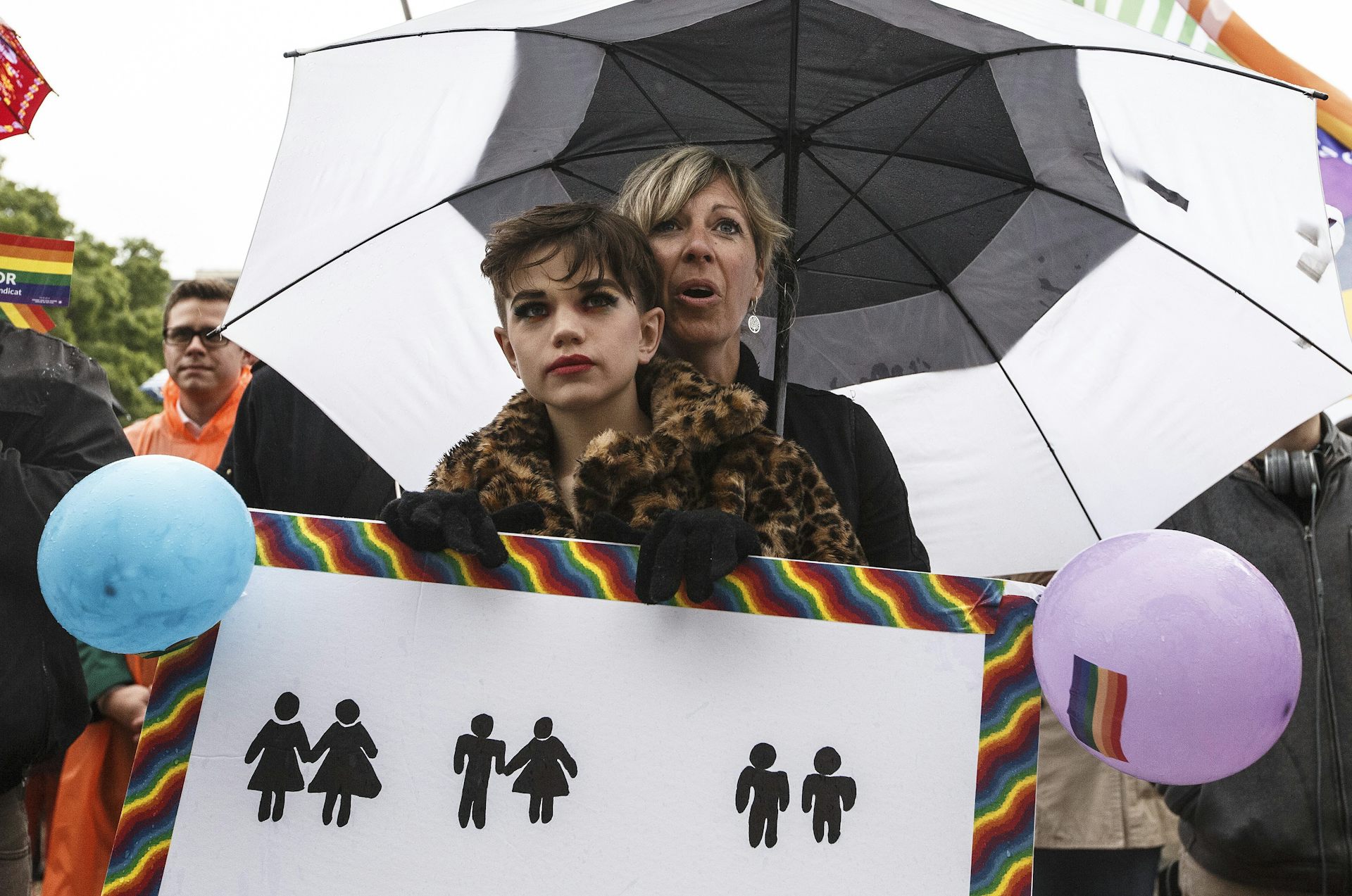 Un adulto con un paraguas oculta a un joven maquillado y sosteniendo un cartel que muestra los íconos de género que se ven comúnmente en los baños, con parejas representadas de mujer-mujer, hombre-mujer y hombre-hombre.