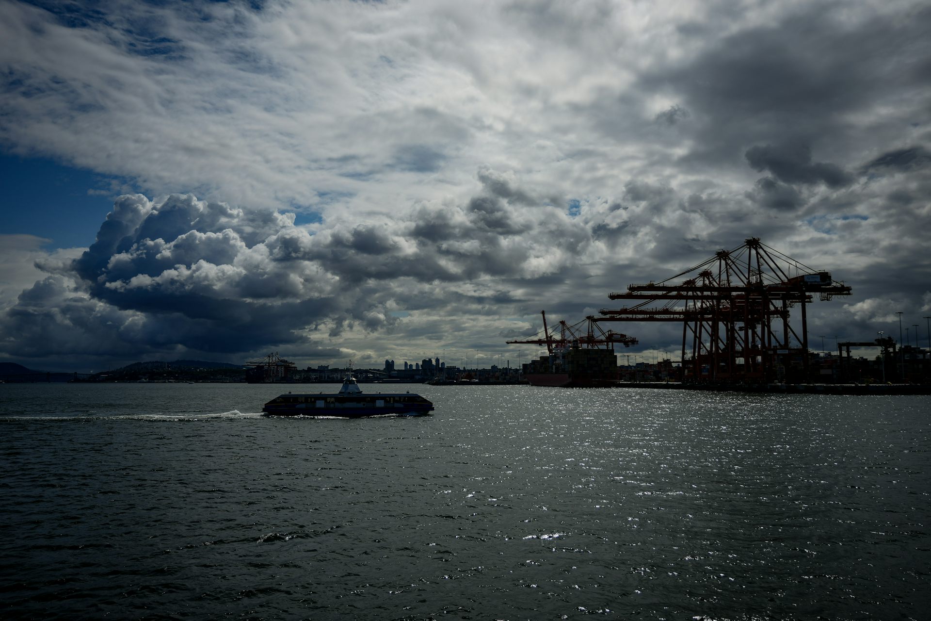an overcast sky above a boat sailing across a body of water with cranes at a port in the background