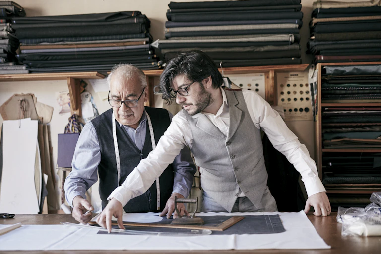 A tailor and a younger assistant, who appears to be his son, measure cloth together.