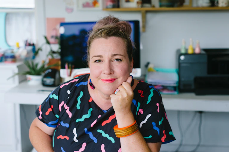A woman smiles softly as she looks straight at the camera, seated and wearing a dark top with colorful squiggles on it.