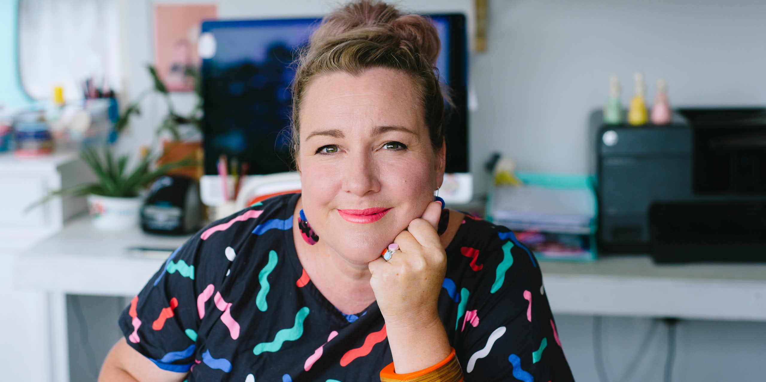 A woman smiles softly as she looks straight at the camera, seated and wearing a dark top with colorful squiggles on it.