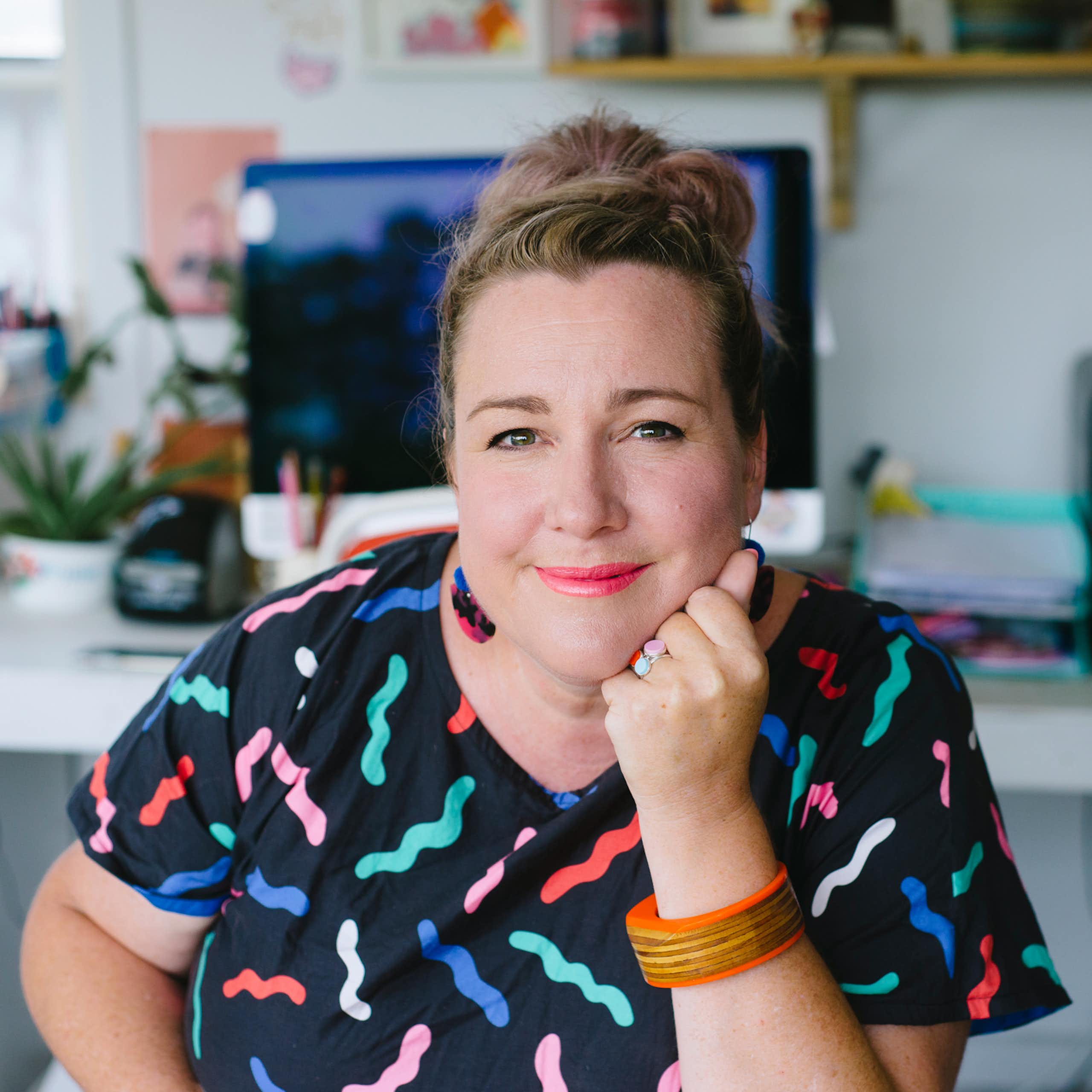 A woman smiles softly as she looks straight at the camera, seated and wearing a dark top with colorful squiggles on it.