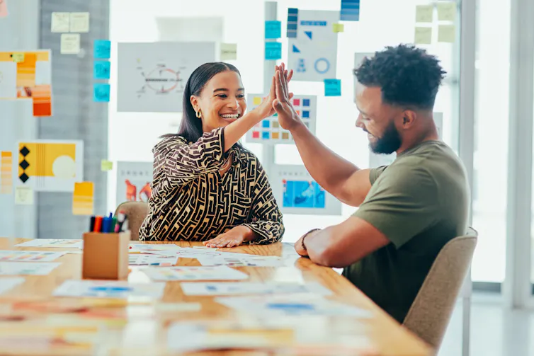 A man and woman seated at an office table high-five each other in a room whose glass walls are covered with print-outs and sticky notes.