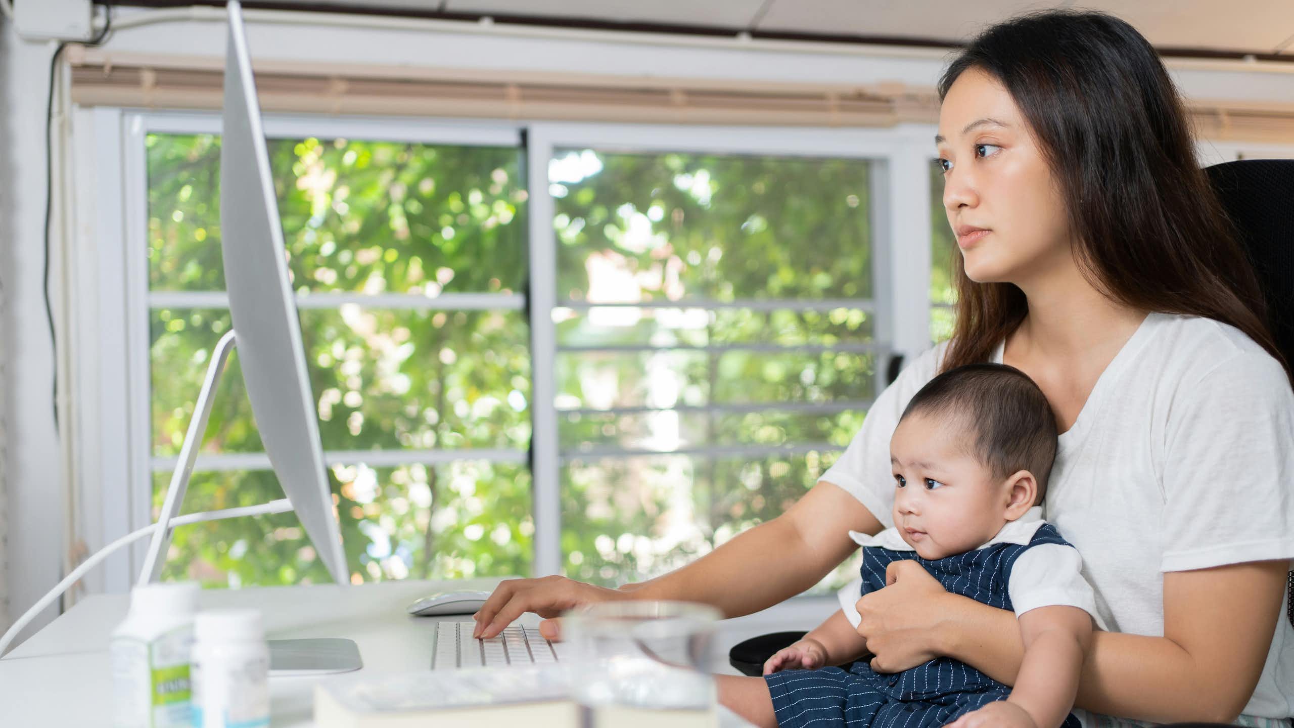 A woman in front of a computer and holding her baby.
