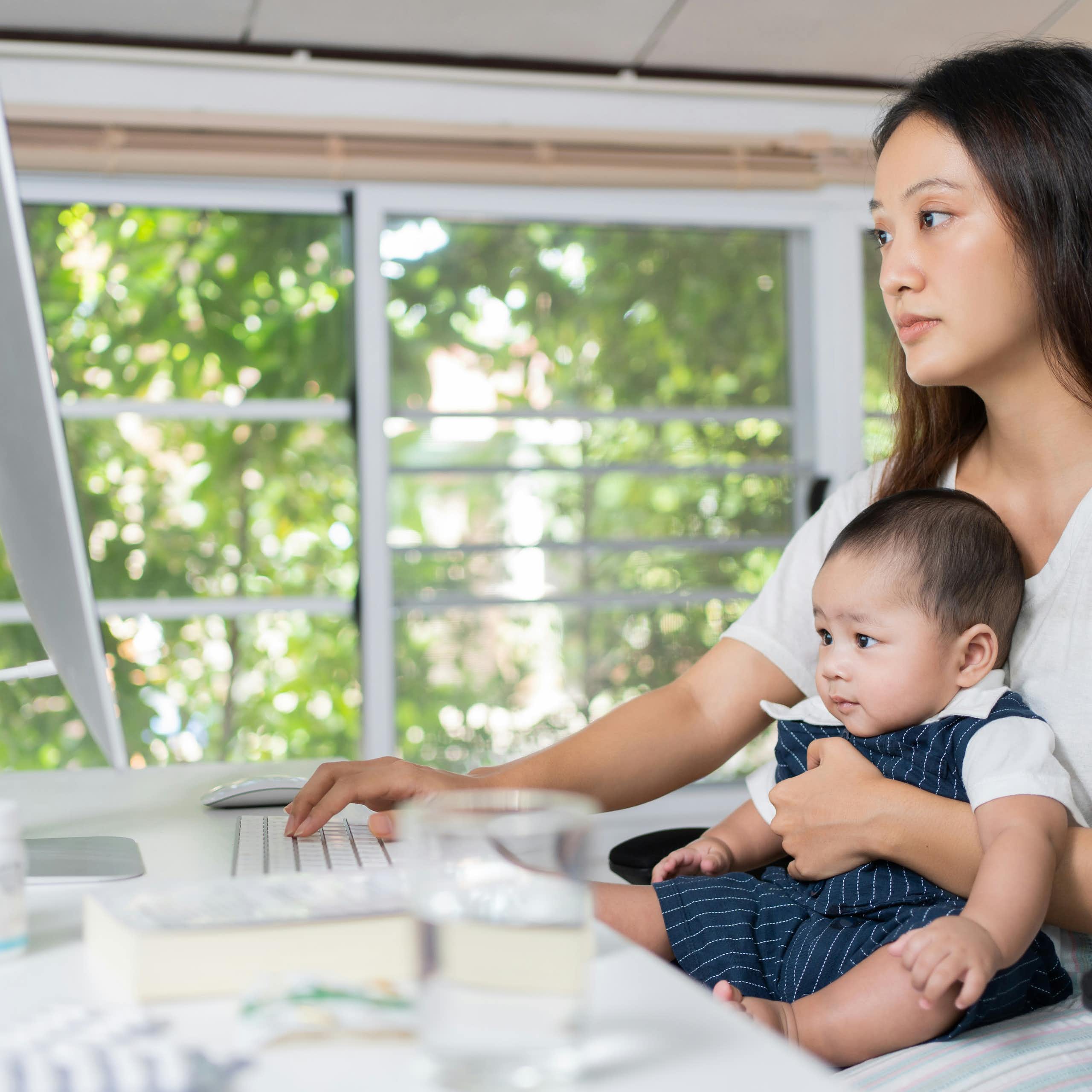 A woman in front of a computer and holding her baby.