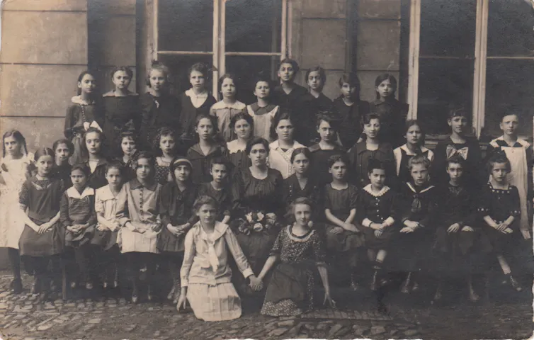 A black and white photo shows several rows of girls formally posed for a large class picture outside.