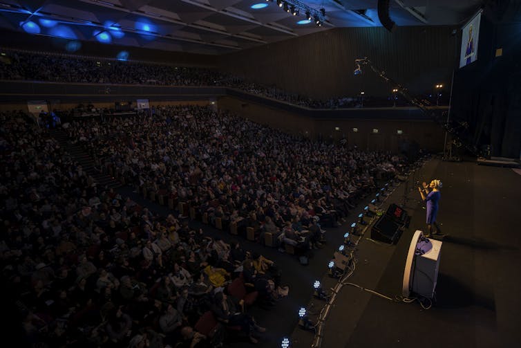 Orthodox Judaism is making area for girls’s spiritual management – even with out conventional ordination 1 A woman in a purple dress and headwrap gestures as she speaks on stage, facing a large, darkened auditorium full of people.