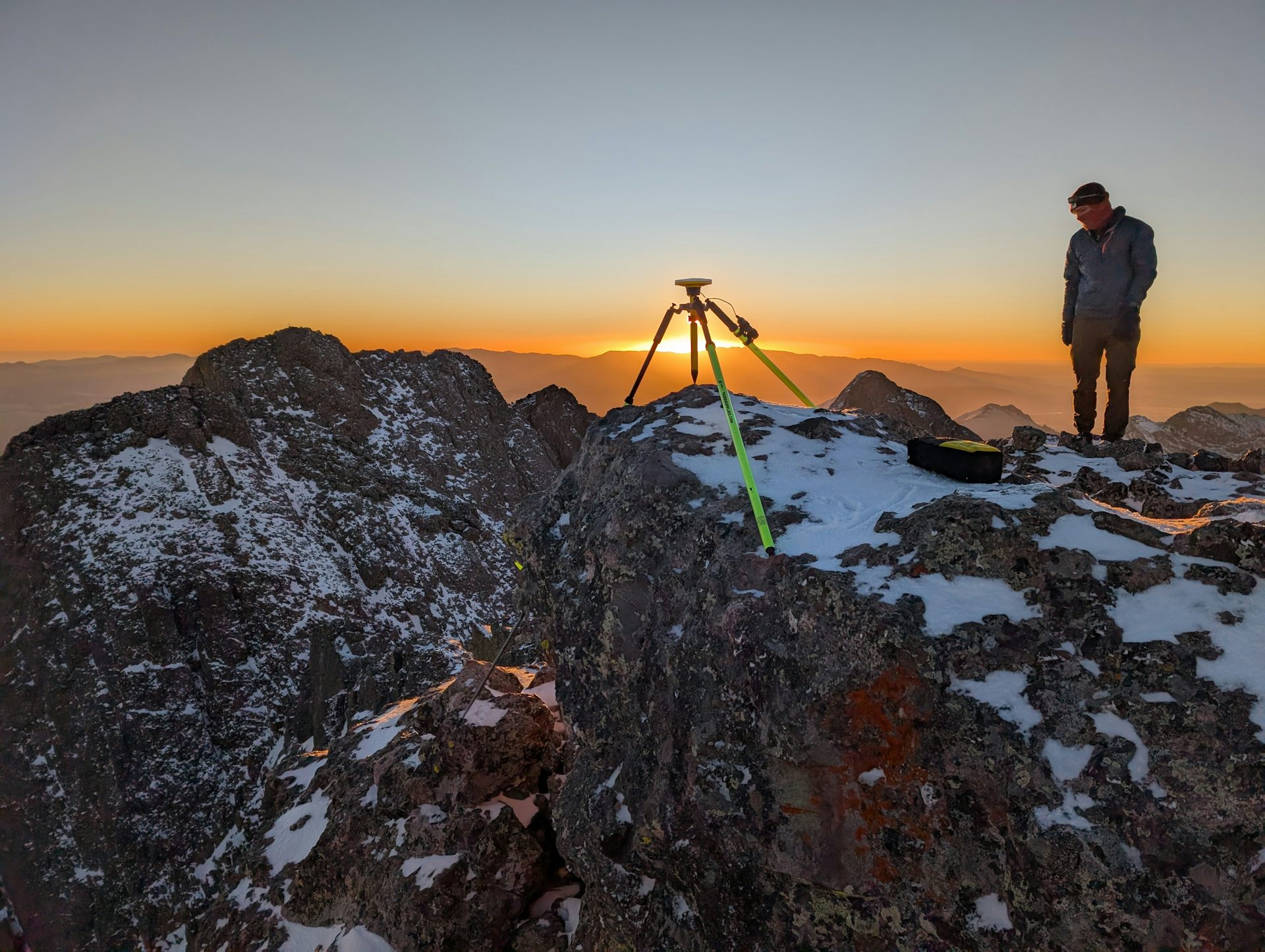 A man stands near a tripod at the top of a mountain.