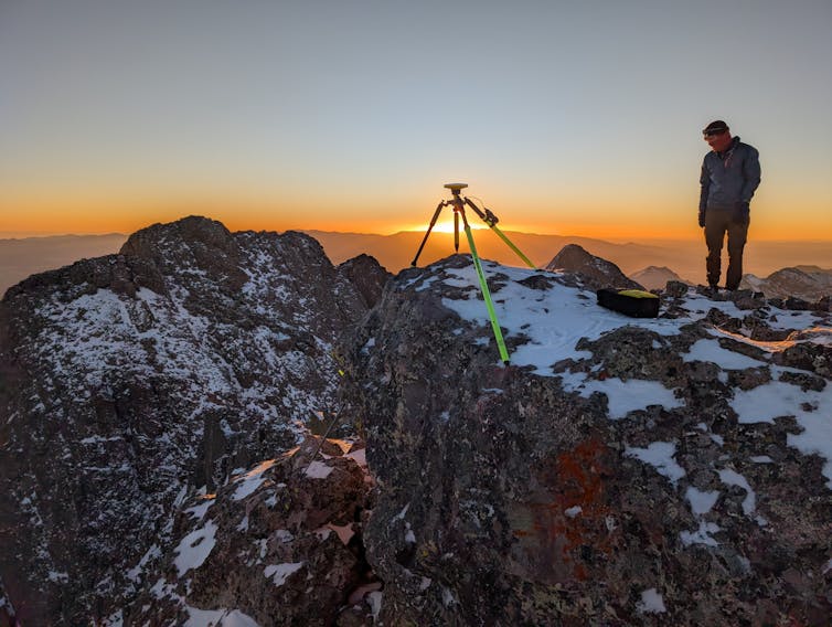 A man stands near a tripod at the top of a mountain.