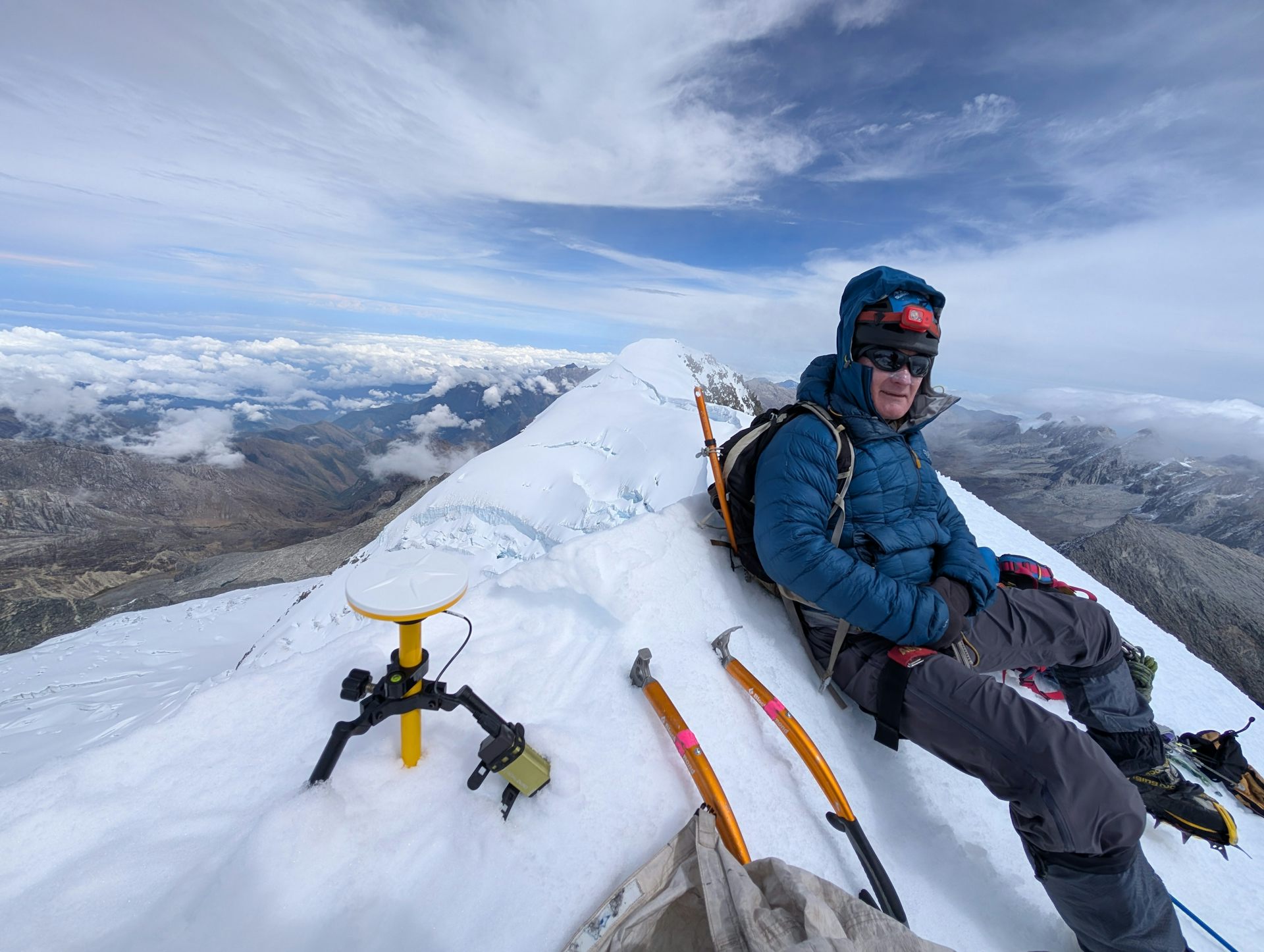 A man in cold-weather clothing sits on an icy mountain peak.