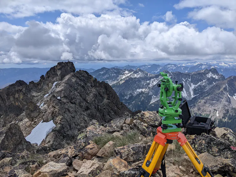 A greean machine with yellow tripod legs sits on a mountain top.
