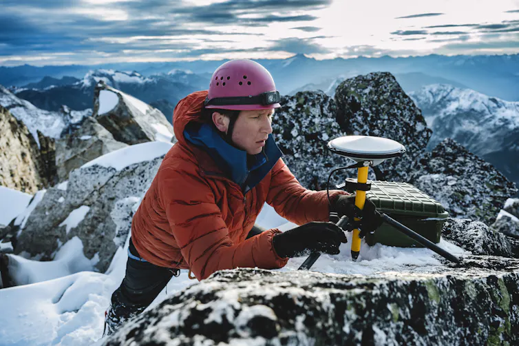 A man with a red helmet and jacket adjusts a small device on the top of a mountain.