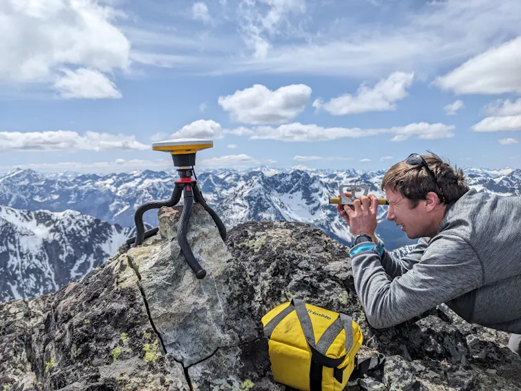 A small flat instrument sits on a tripod near a man who holds a measuring device to his eyes.