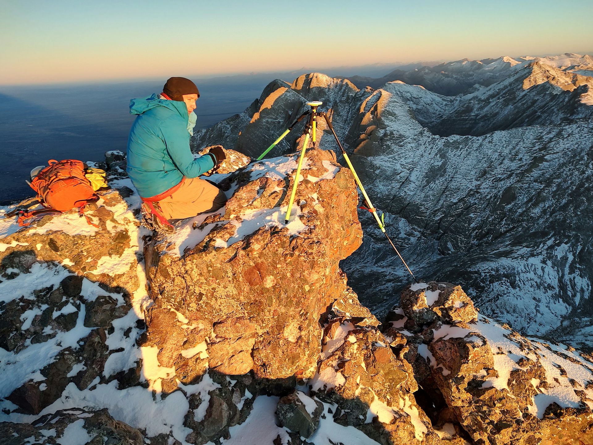 A man in a green jacket sits near a device on a tripod on the top of a mountain.