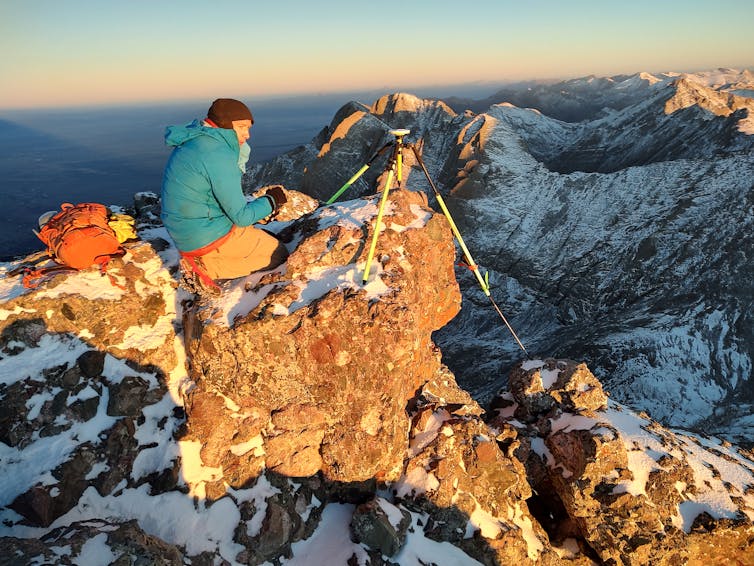 Measuring Colorado’s mountains one hike at a time 6 A man in a green jacket sits near a device on a tripod on the top of a mountain.