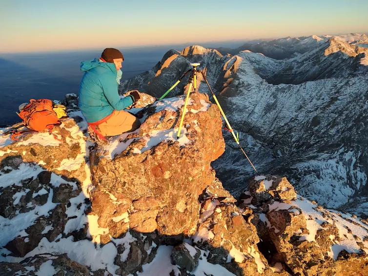 A man in a green jacket sits near a device on a tripod on the top of a mountain.