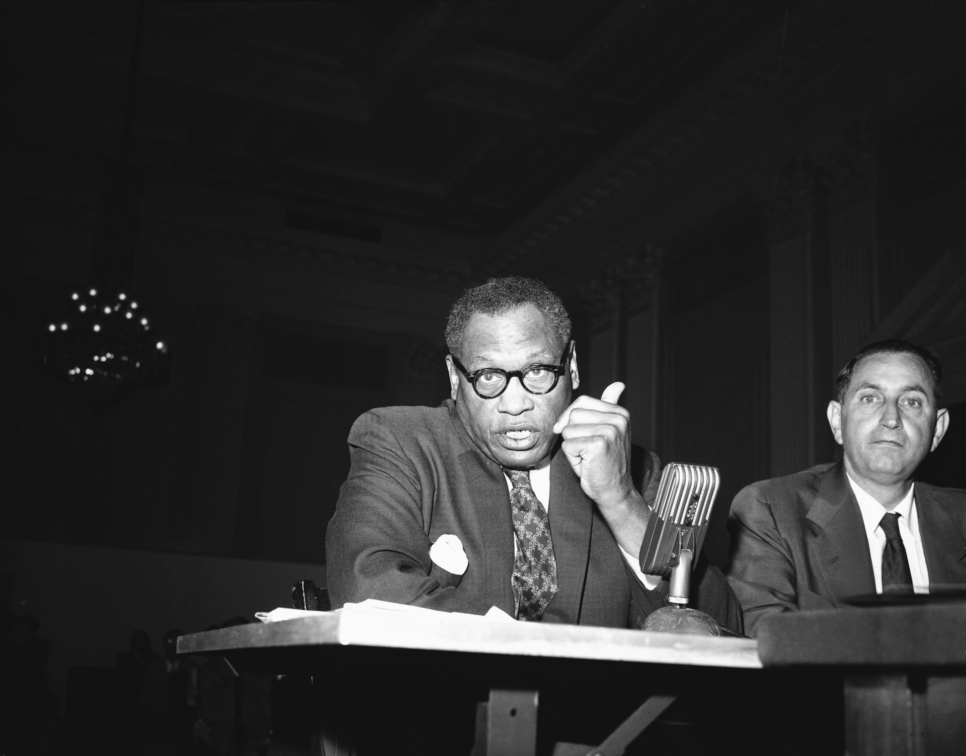 A Black man sits in front of a table with a microphone on it.