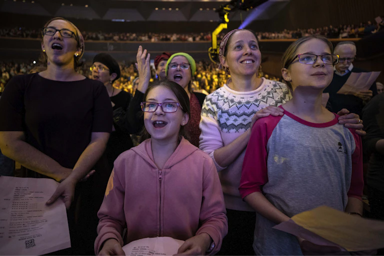 Two women and two girls smile and seem to chant or sing as they stand in a large crowd of women in an auditorium.