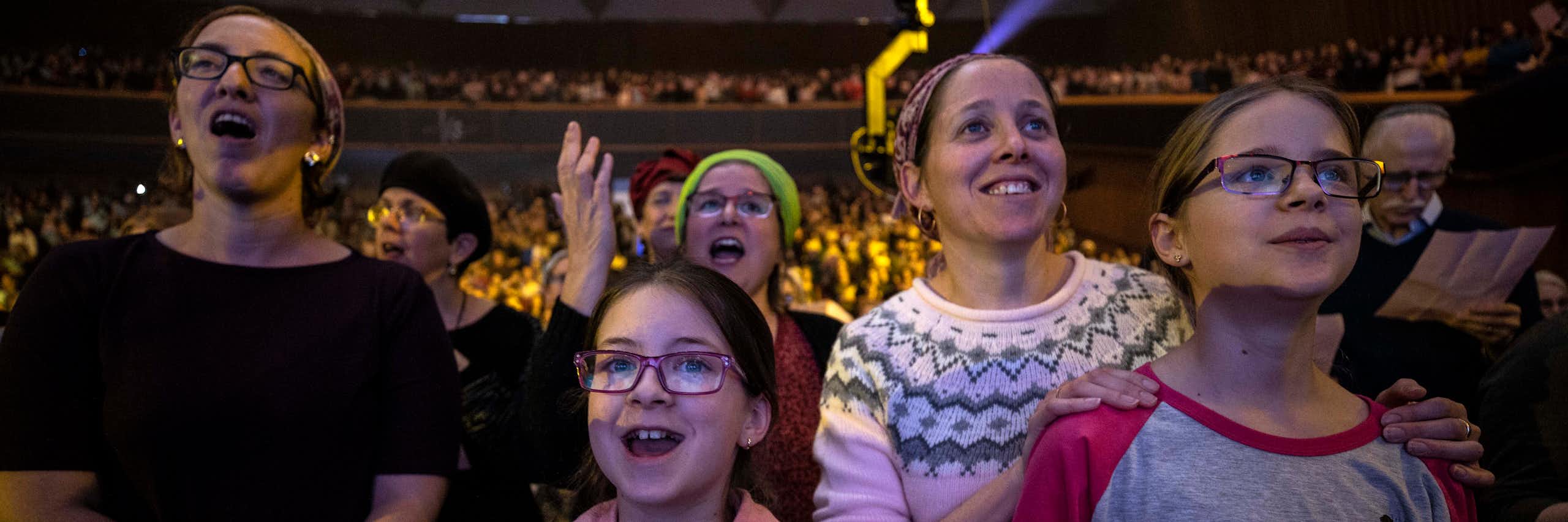 Two women and two girls smile and seem to chant or sing as they stand in a large crowd of women in an auditorium.