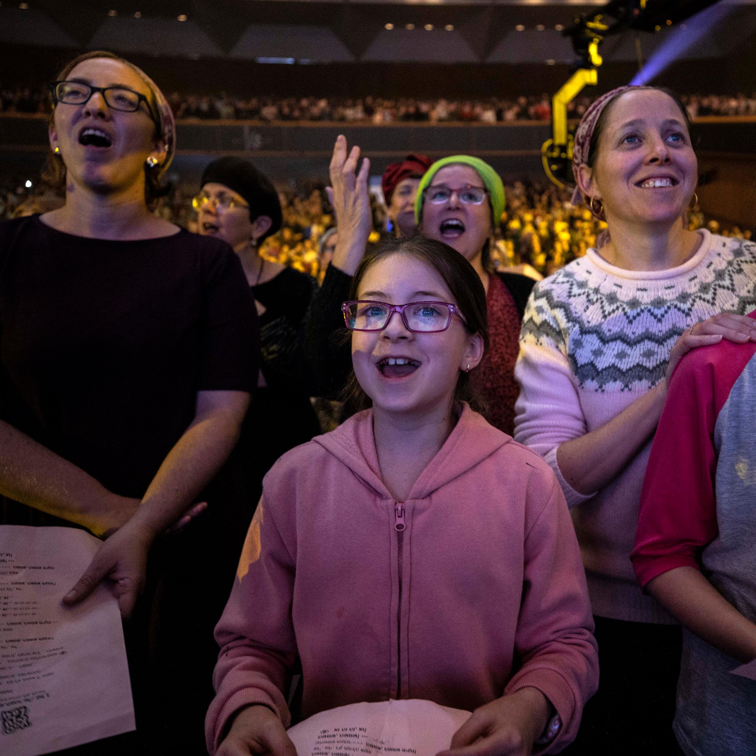 Two women and two girls smile and seem to chant or sing as they stand in a large crowd of women in an auditorium.