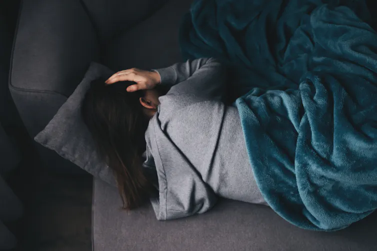 Person curled up on couch beneath a blanket, hand over head