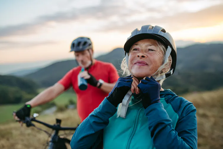 A smiling woman adjusts her bicycle helmet while a man on a bike sips from a water bottle in the background