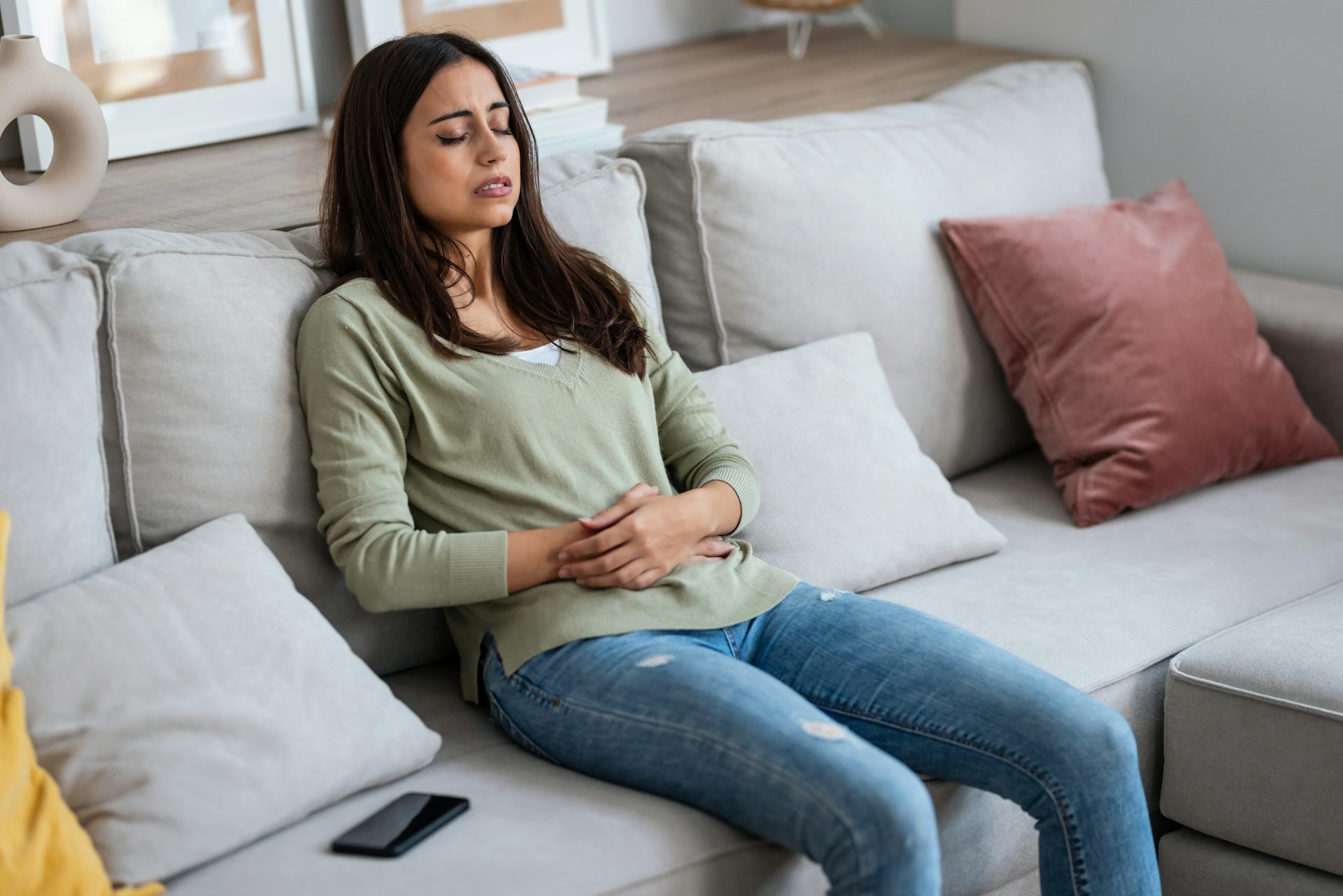 Une jeune femme se tient le ventre, assise sur un canapé, les yeux fermés.