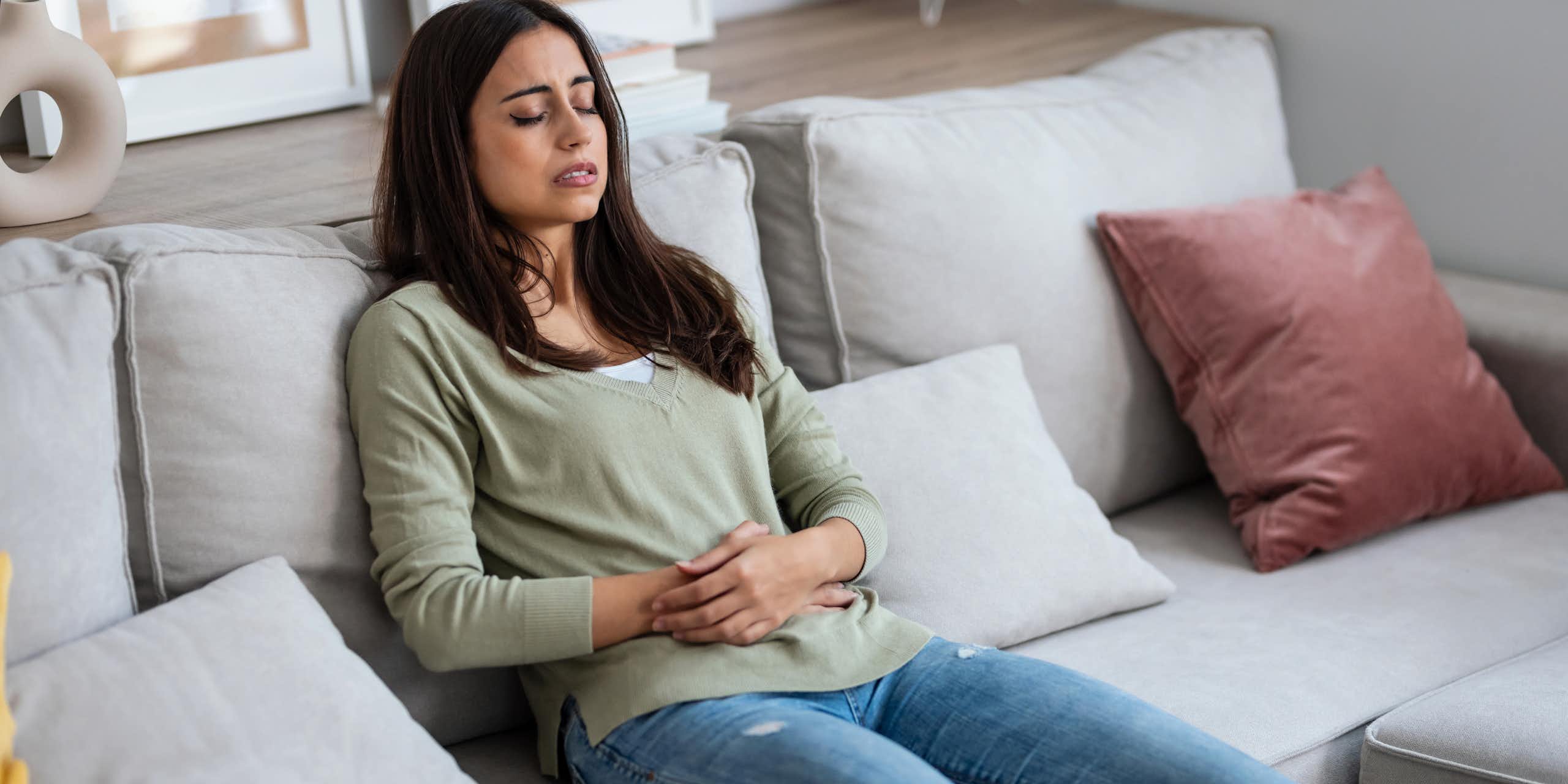 Une jeune femme se tient le ventre, assise sur un canapé, les yeux fermés.