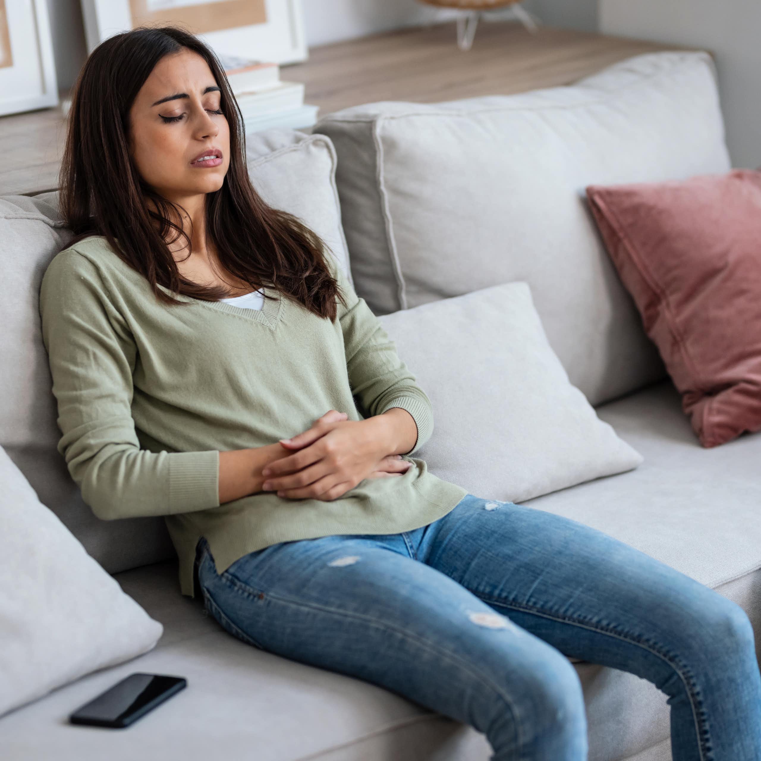 Une jeune femme se tient le ventre, assise sur un canapé, les yeux fermés.