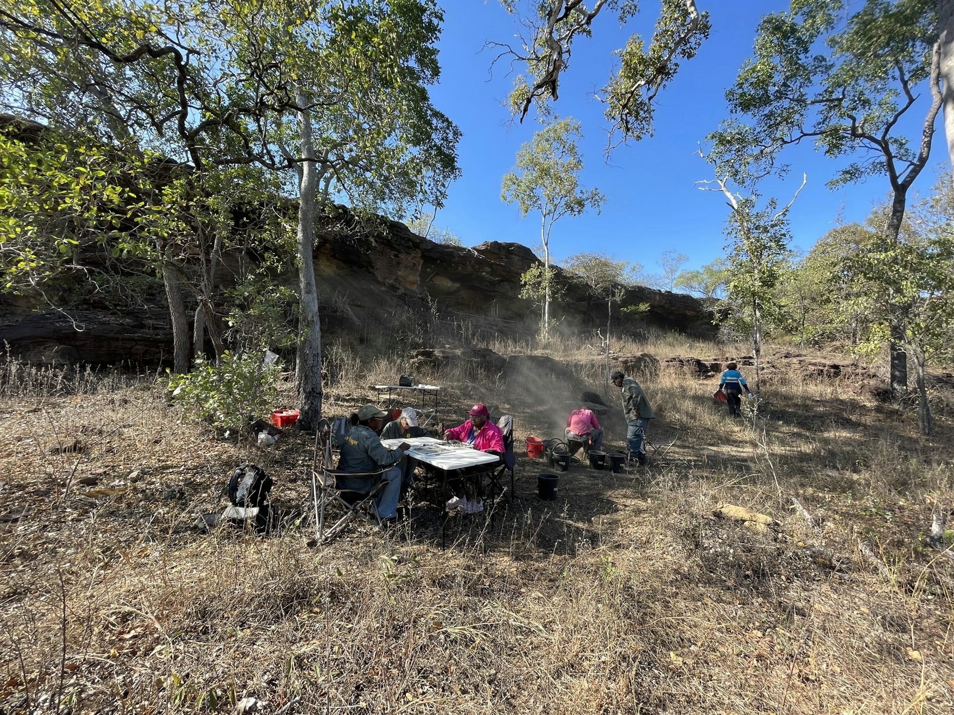 People sitting around a table in the shade sorting archaeological materials, in front of the sandstone rock shelter site that the materials have been excavated from.