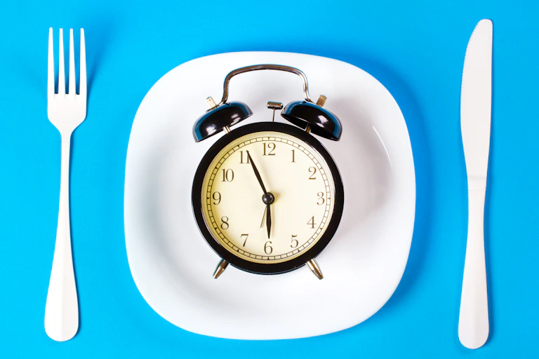 A black clock sits on a dinner plate, with a fork and knife on either side. It is almost 6 o'clock.