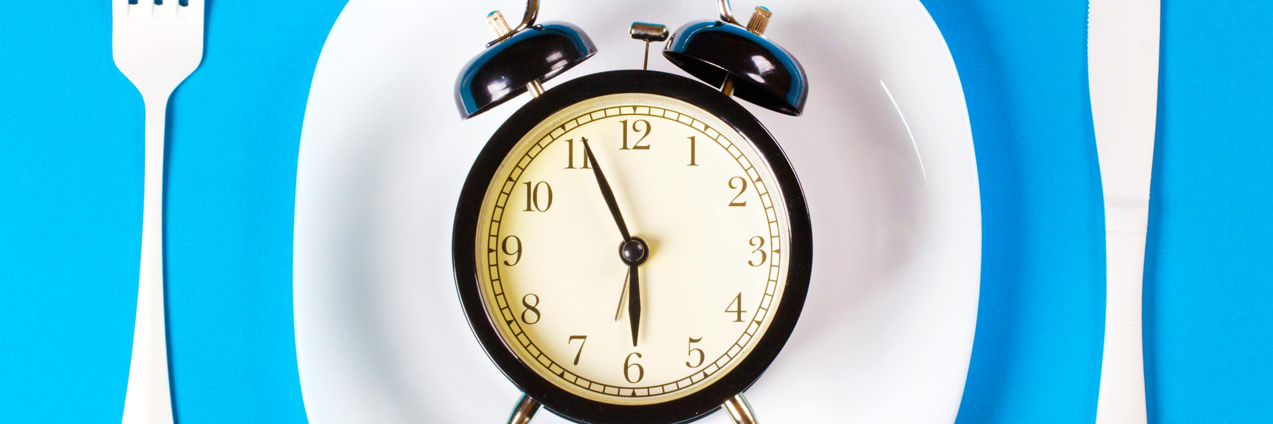 A black clock sits on a dinner plate, with a fork and knife on either side. It is almost 6 o'clock.