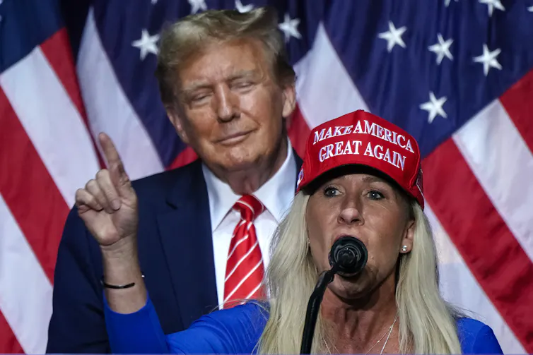 Une femme blonde coiffée d’un bonnet rouge parle devant un micro tandis qu’un homme en costume se tient derrière elle, avec des drapeaux américains en arrière-plan