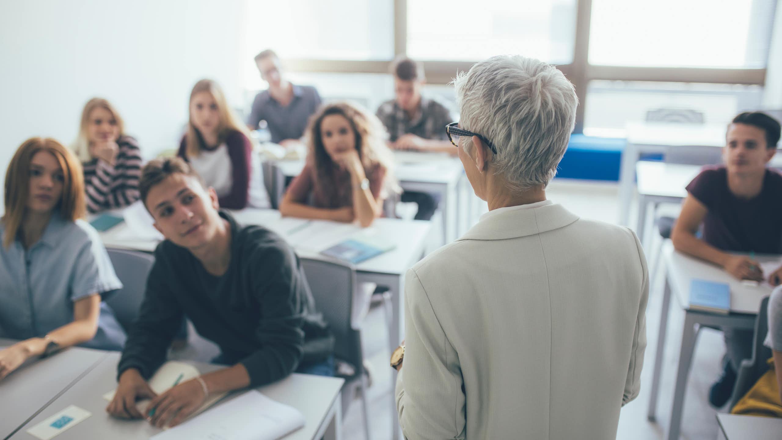 una mujer de espaldas habla a los alumnos de una clase.