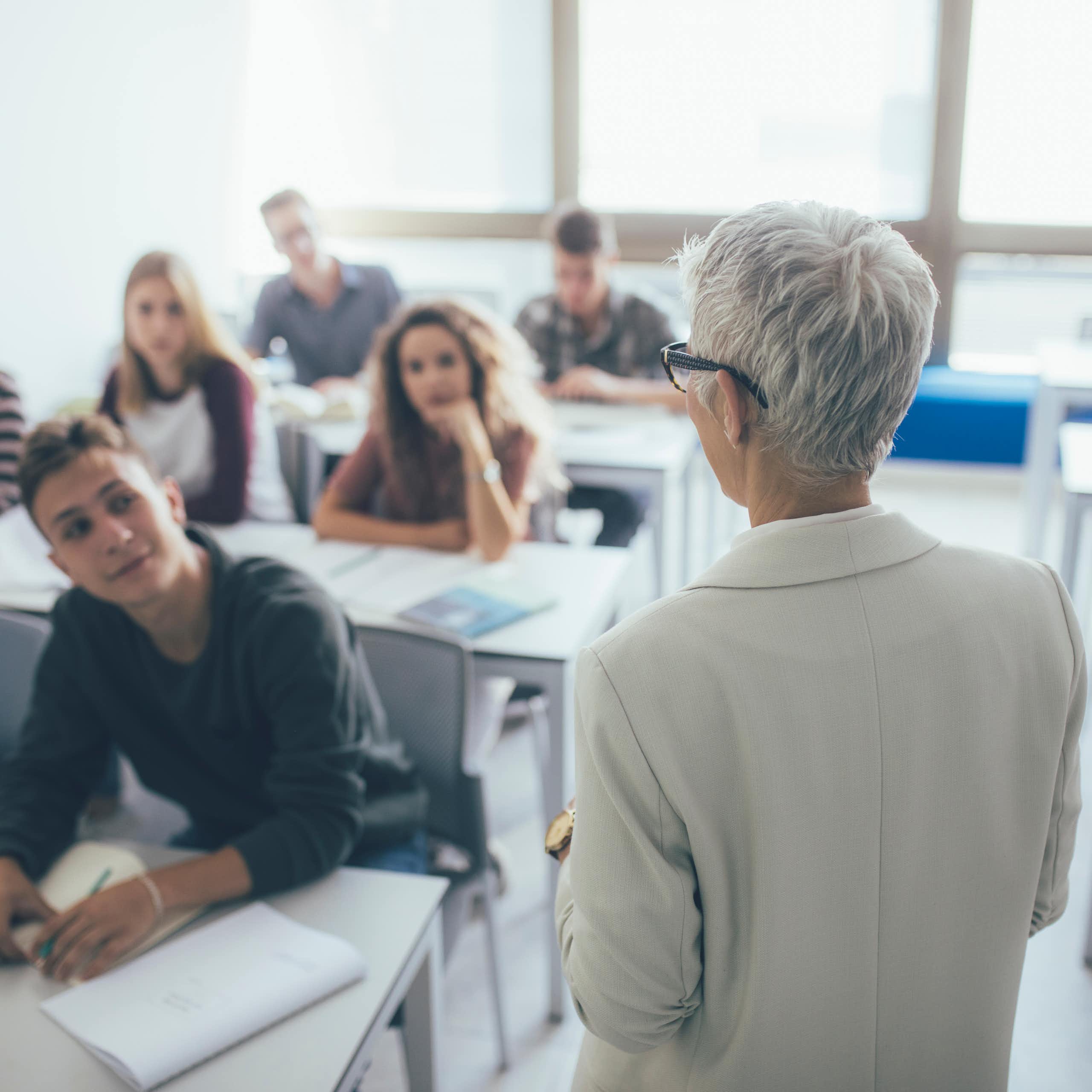 una mujer de espaldas habla a los alumnos de una clase.