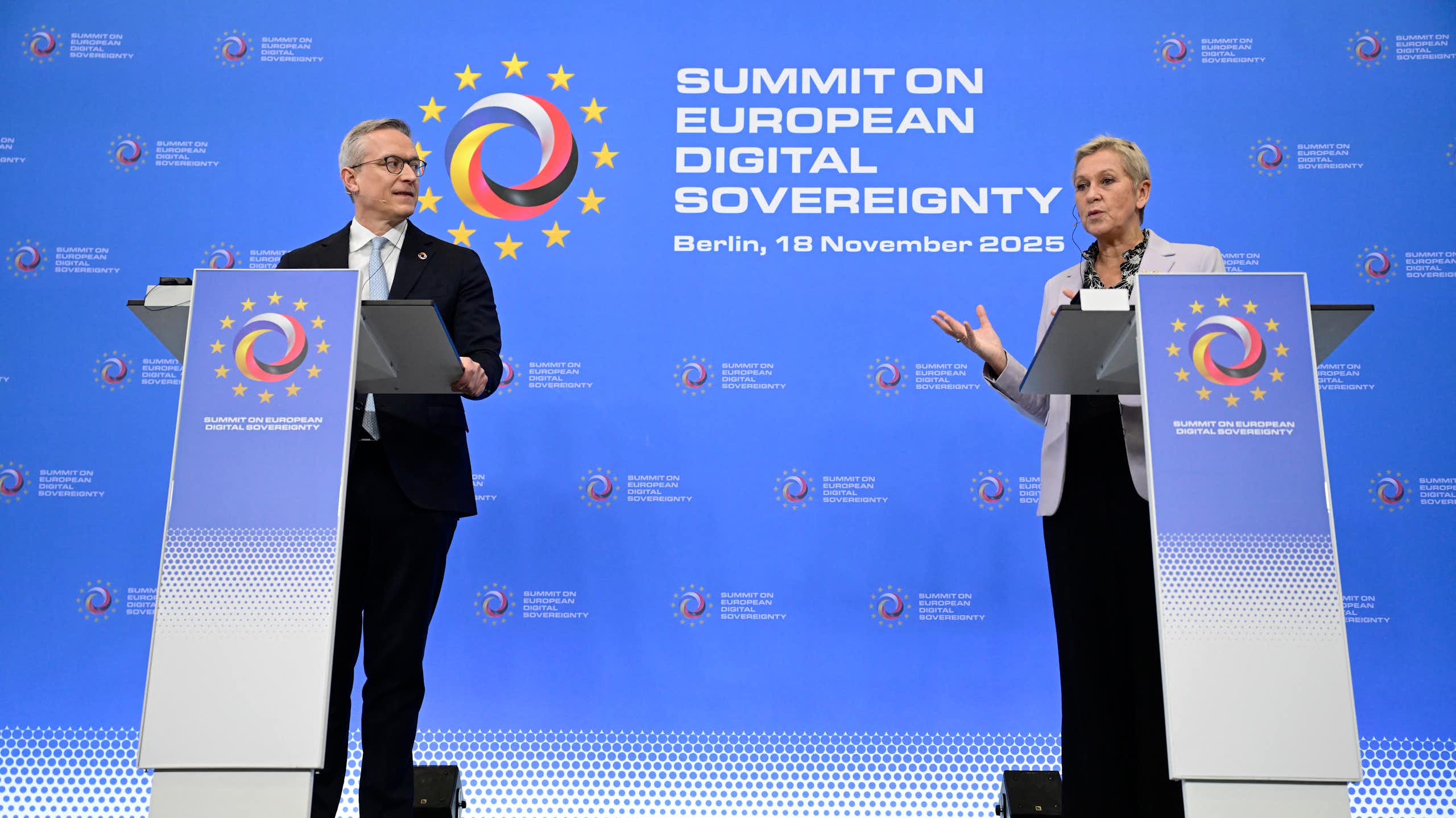 A man and a woman stand behind lecterns on a stage. The woman is speaking.