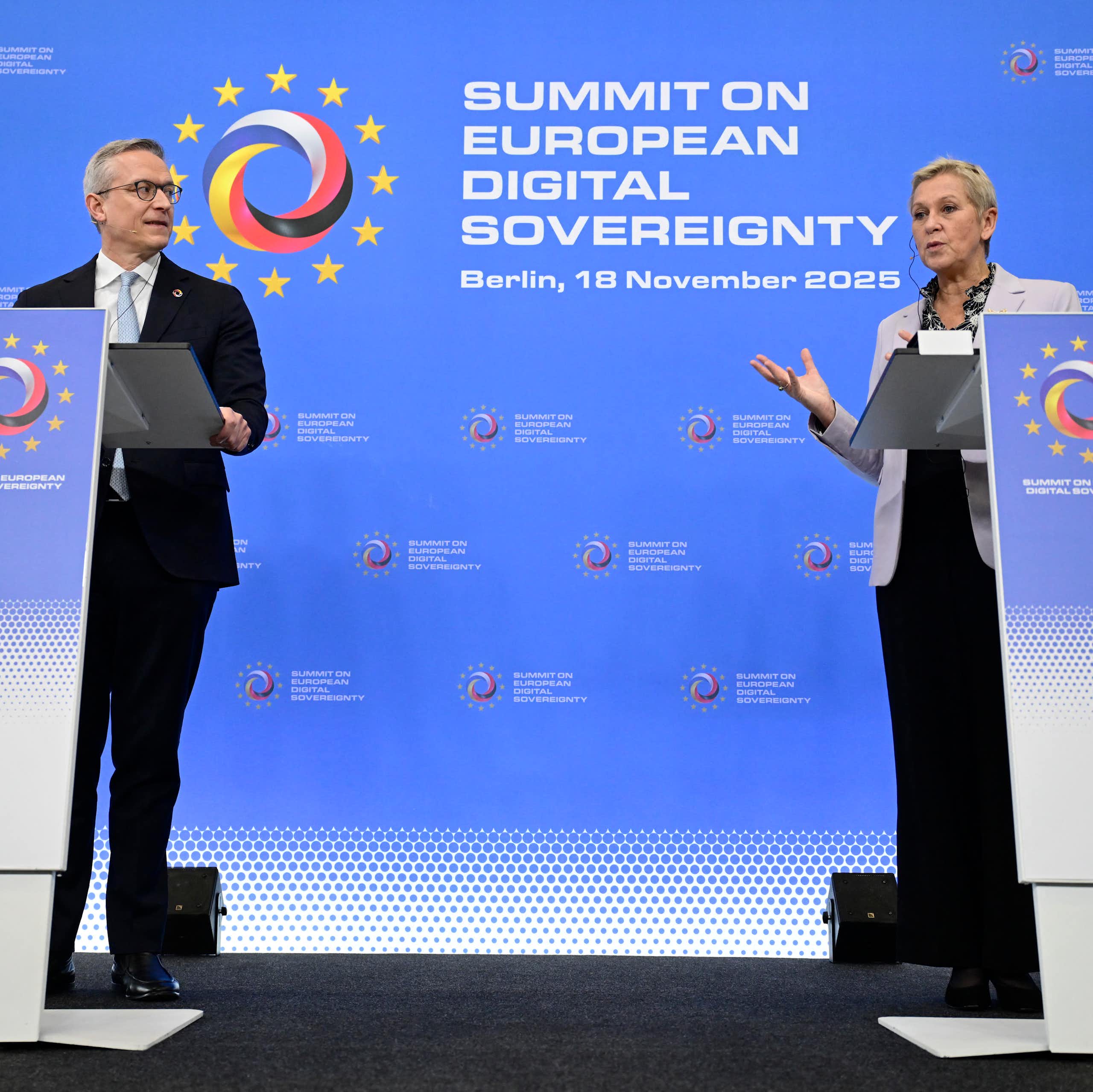 A man and a woman stand behind lecterns on a stage. The woman is speaking.