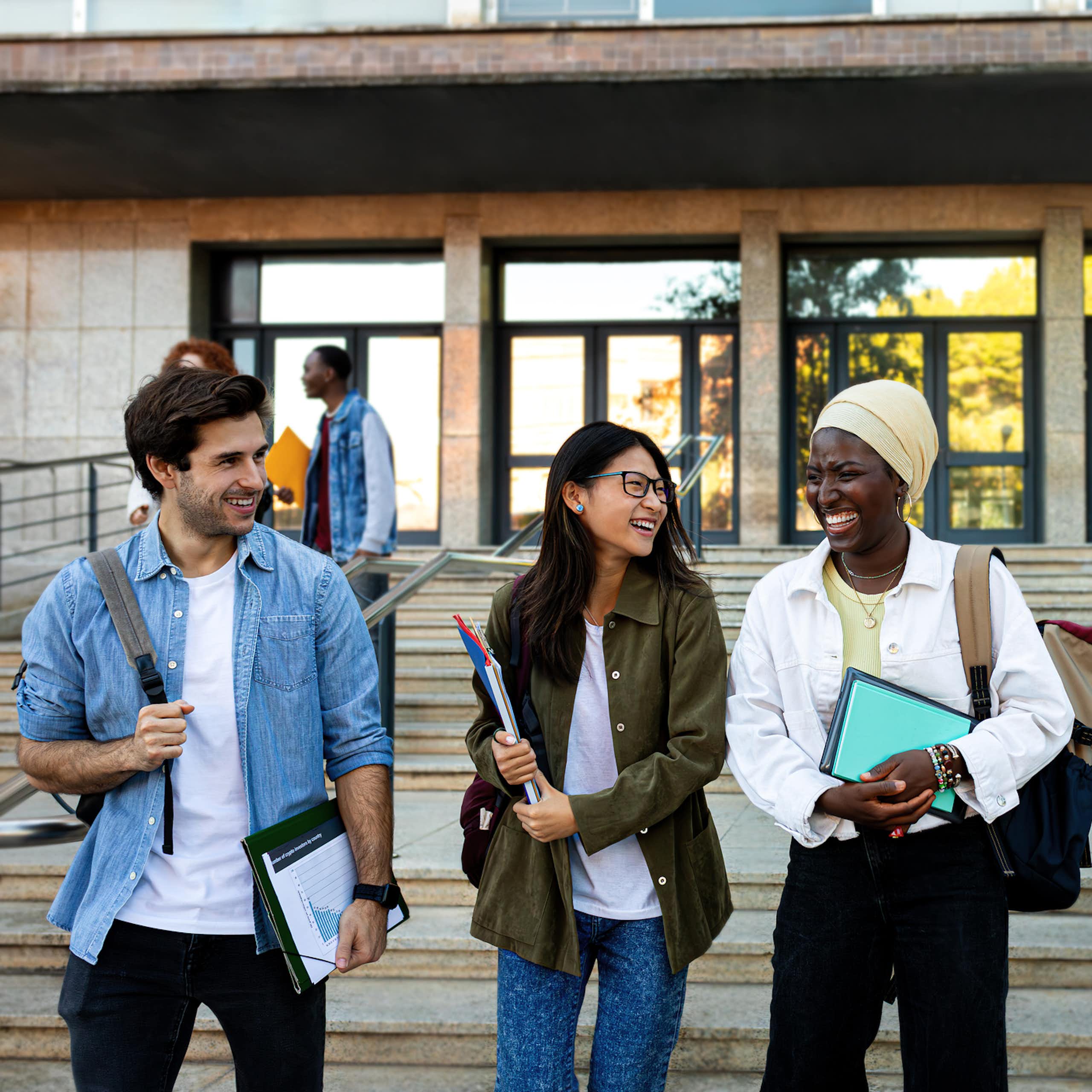 Three college-age students walk down steps outside, smiling and chatting, as other students walk in the background.