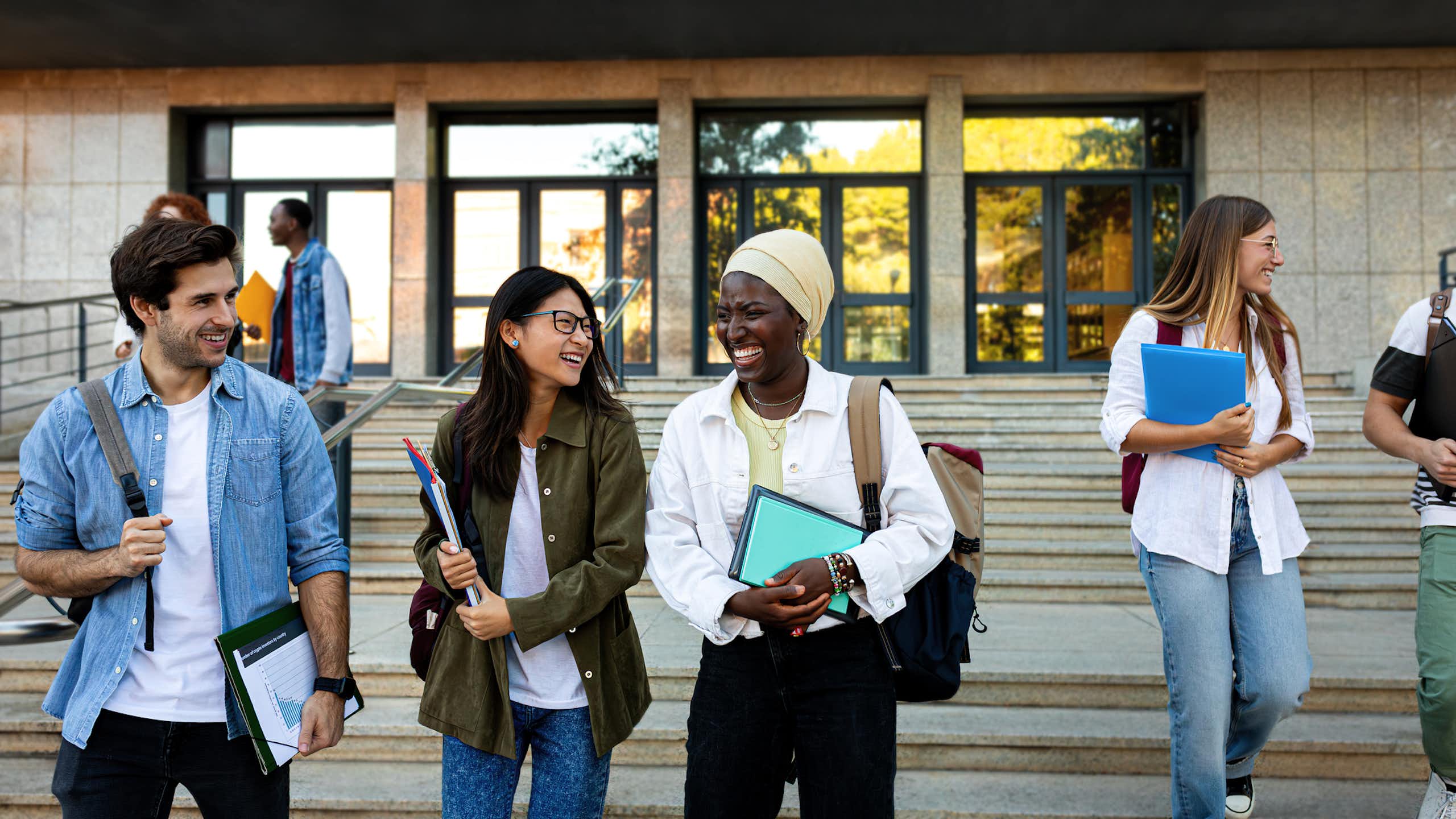 Three college-age students walk down steps outside, smiling and chatting, as other students walk in the background.