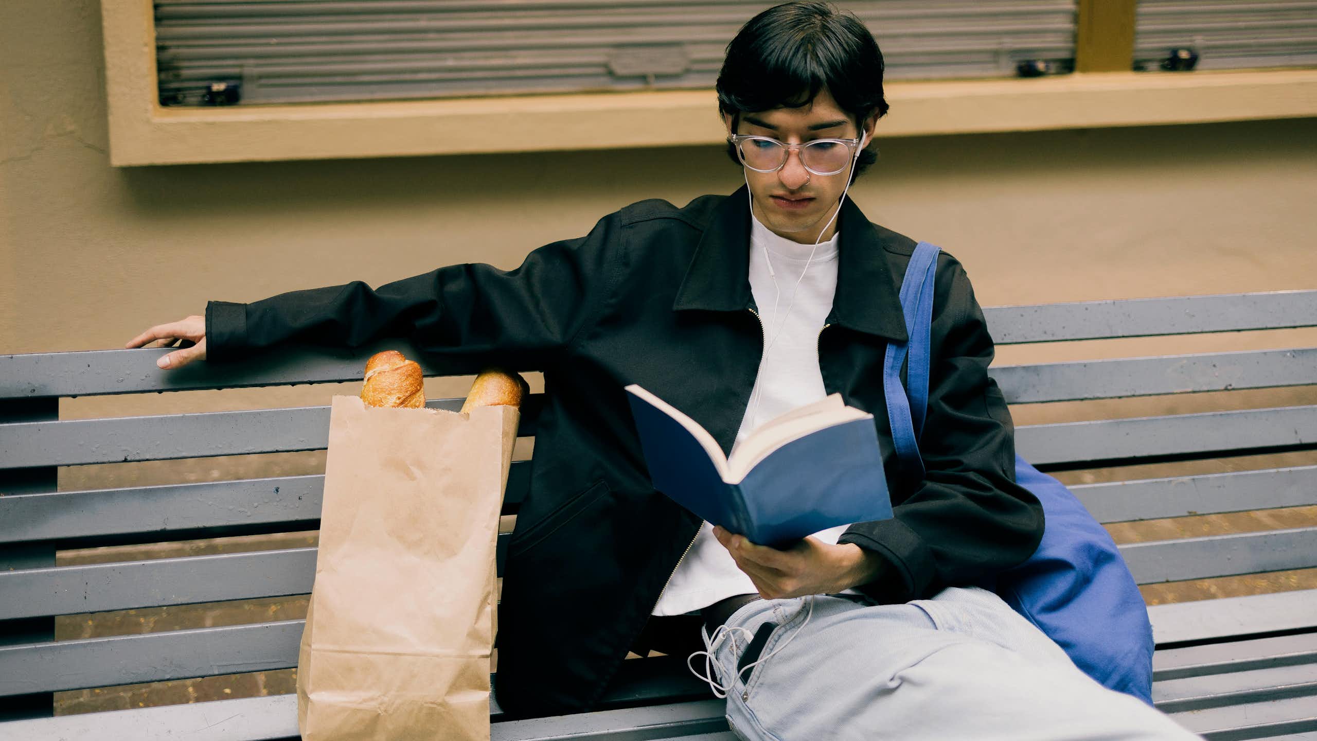 A young man reading a book on a bench with a tote bag slung over his shoulder.
